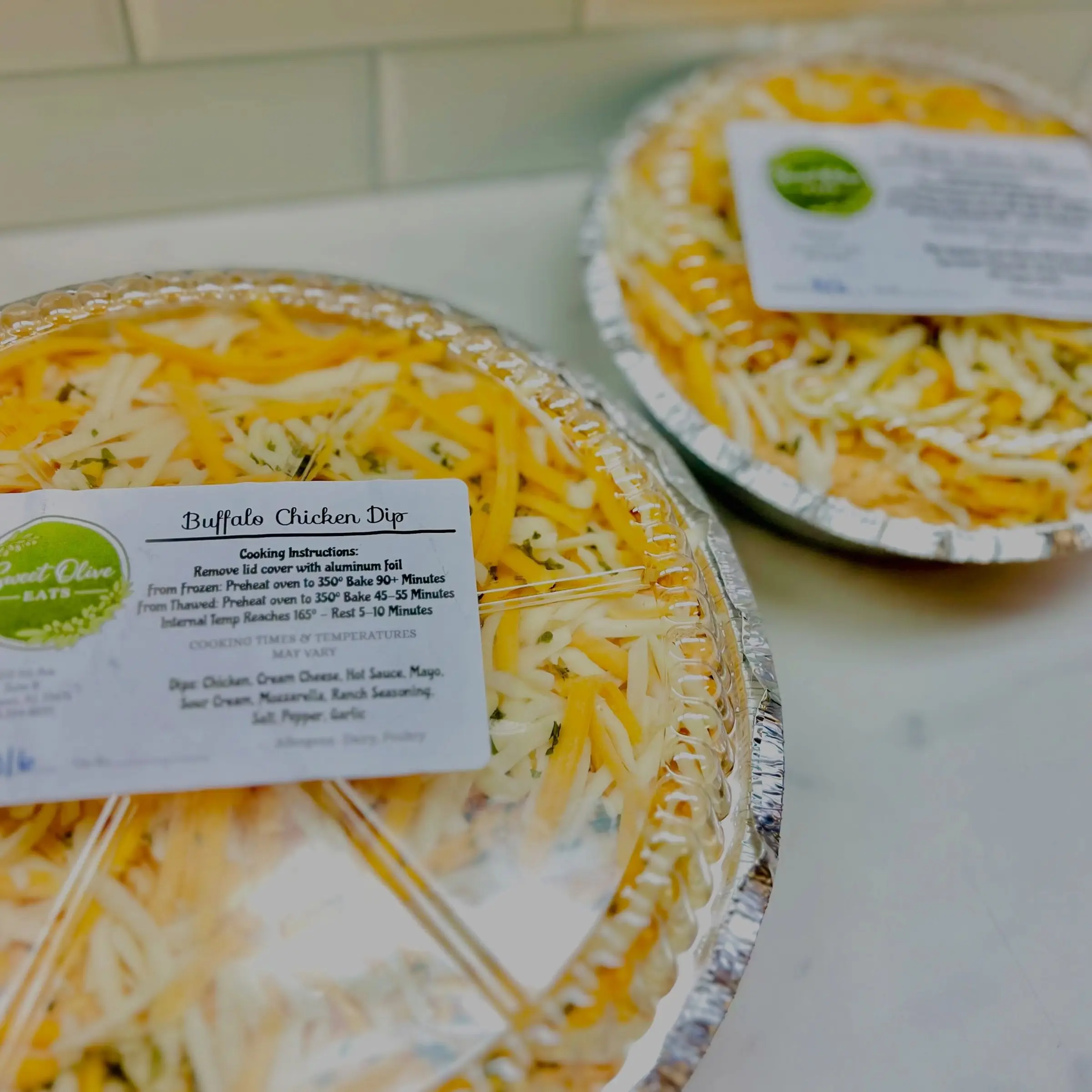 Two Buffalo Chicken Dip pans under clear lids, each with a Sweet Olive label printed with the cooking instructions
