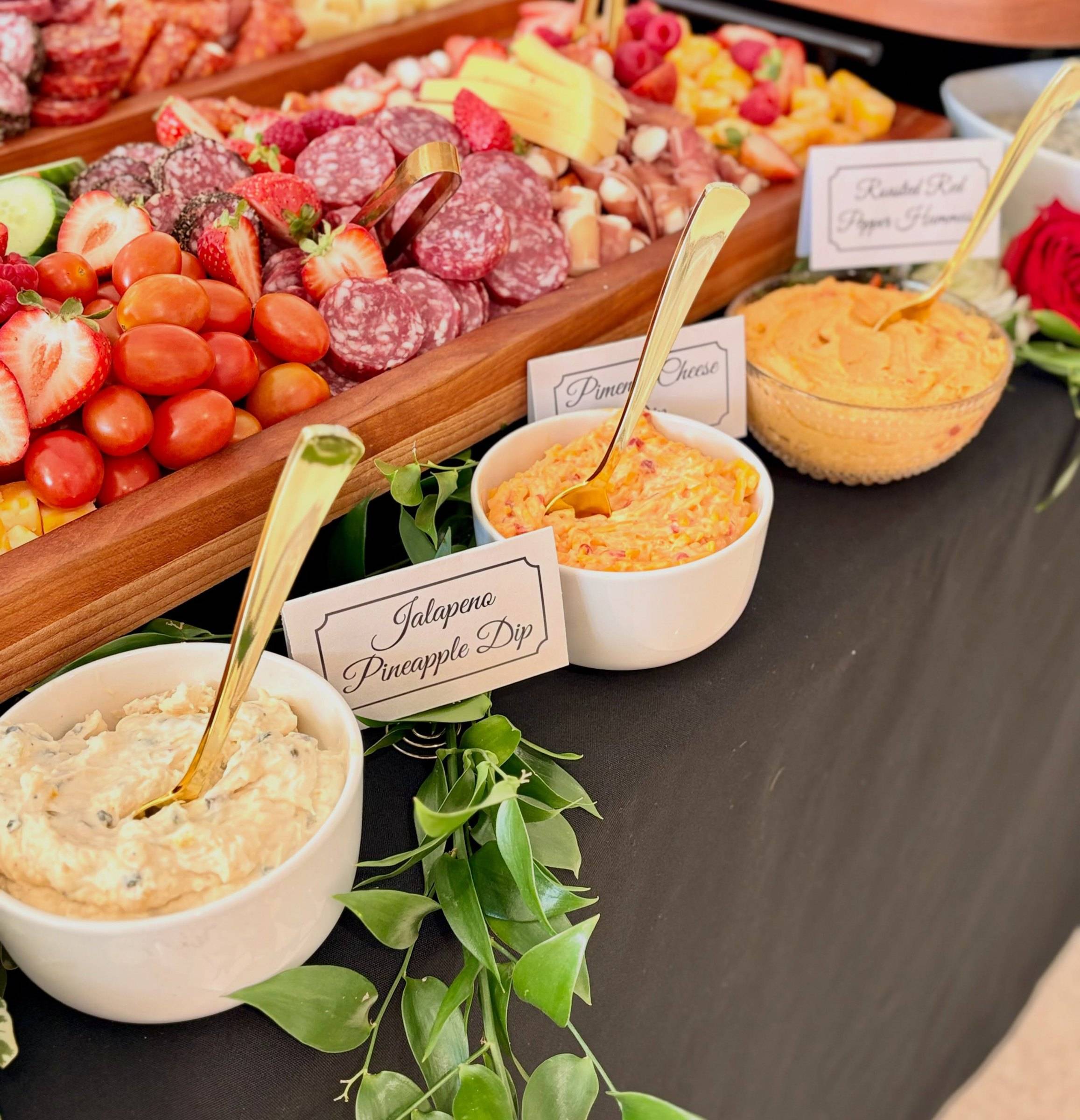 A charcuterie board of salami, tomatoes and strawberries beside bowls of dip with gold spoons and white tent cards