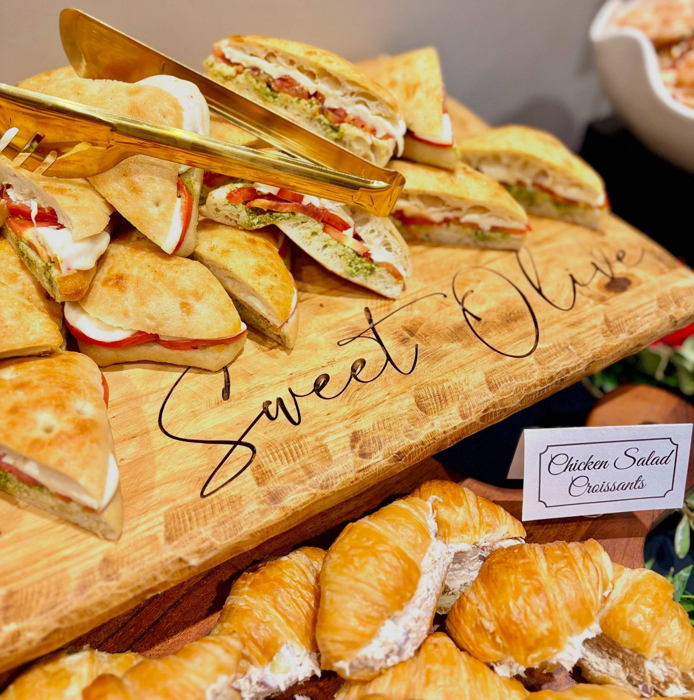 Chicken salad croissants and sandwich halves on a wooden board engraved Sweet Olive in script, with gold tongs
