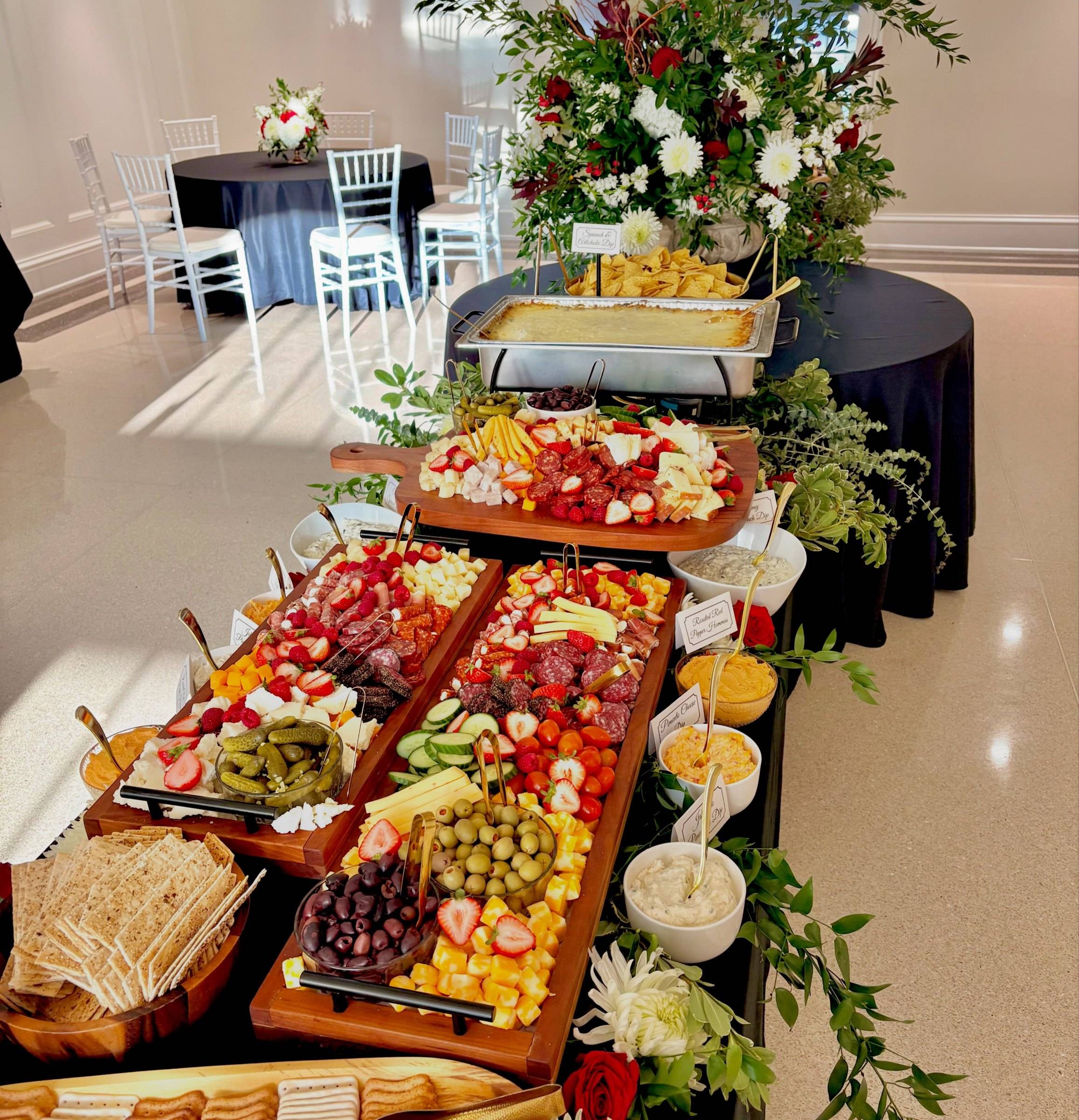 A tiered Sweet Olive grazing spread on black linen in a white event room: trays of salami, cheese, fruit and crackers under a tall arrangement of red and white flowers and greenery
