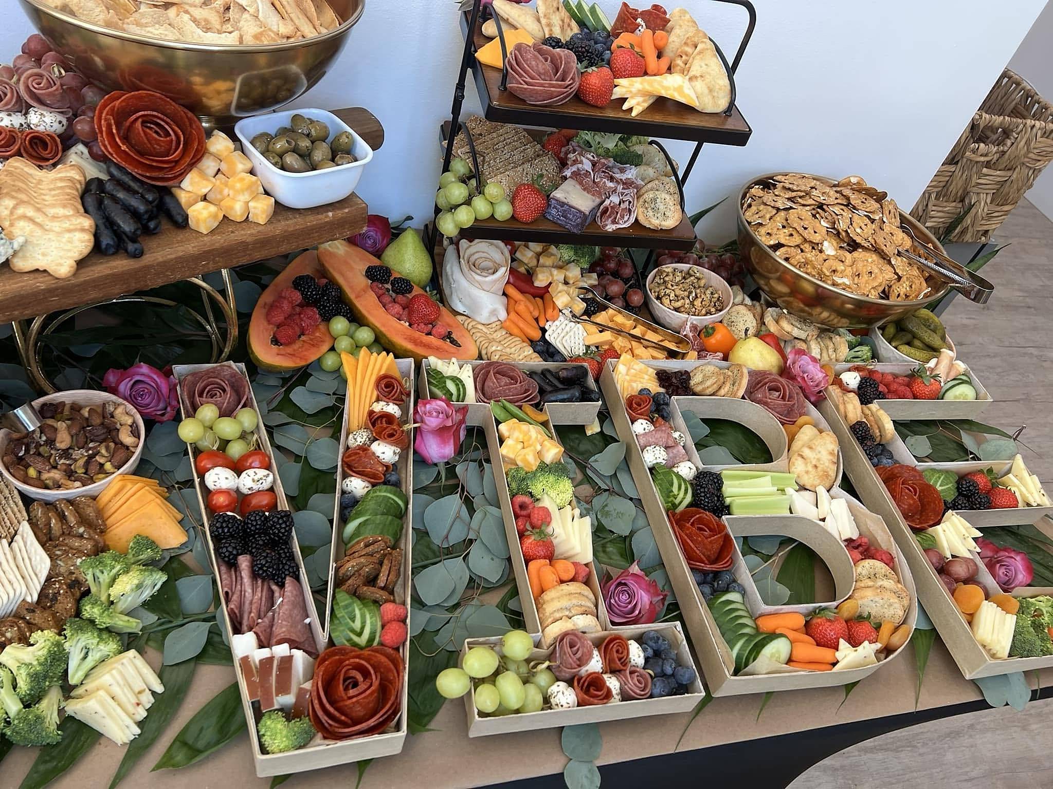 A Sweet Olive grazing table: letter-shaped trays spelling VIBE, filled with salami roses, berries, grapes, mozzarella and cucumbers on eucalyptus, with tiered boards and a bowl of pretzel crisps behind