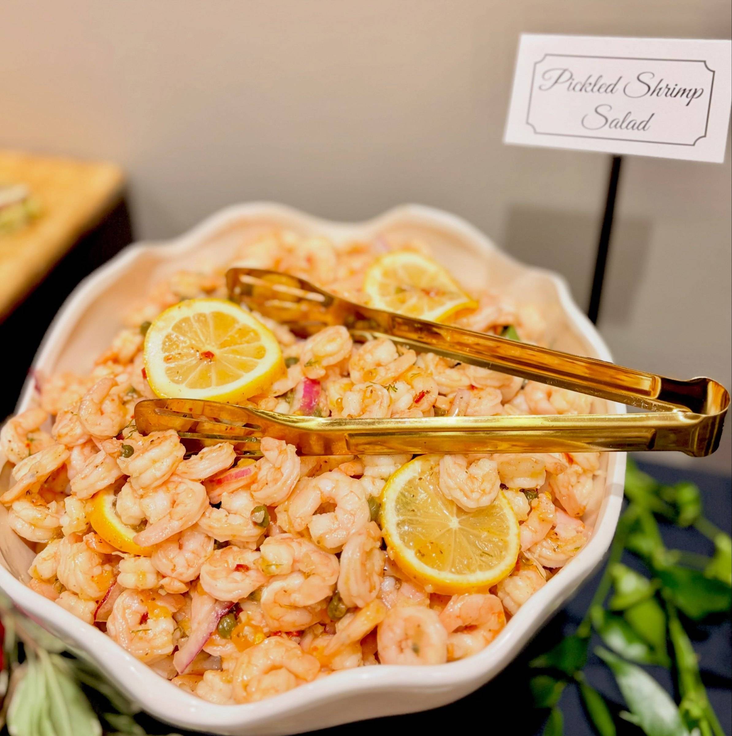 Pickled shrimp salad with lemon slices in a white scalloped bowl, gold tongs resting on top