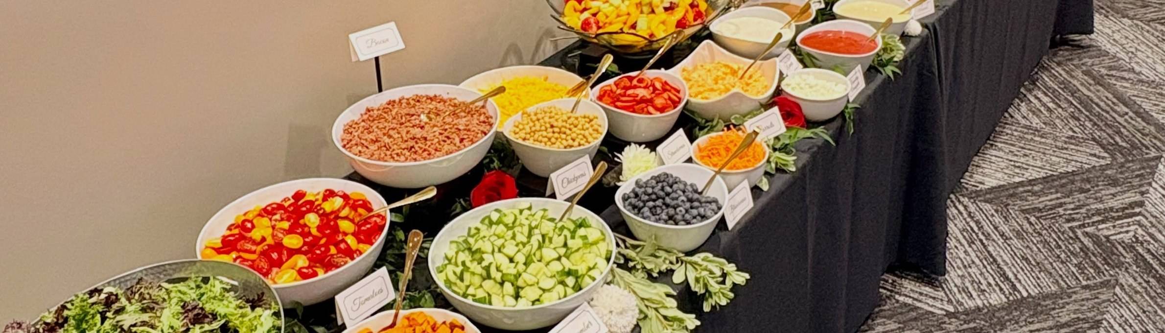 A long Sweet Olive buffet on black linen: white bowls of tomatoes, chickpeas, cucumbers, blueberries and carrots, each with a small white tent card, between red roses and greenery